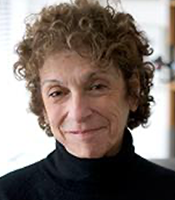 Ann Burack wears a shortly curly pixie cut and a black turtle neck smiling into camera.