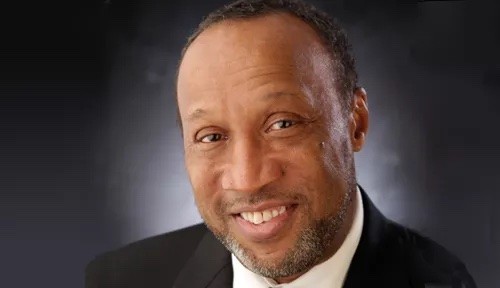 Dr. Lewis, smiles into camera dark background wearing a white button down shirt and a dark blazer.