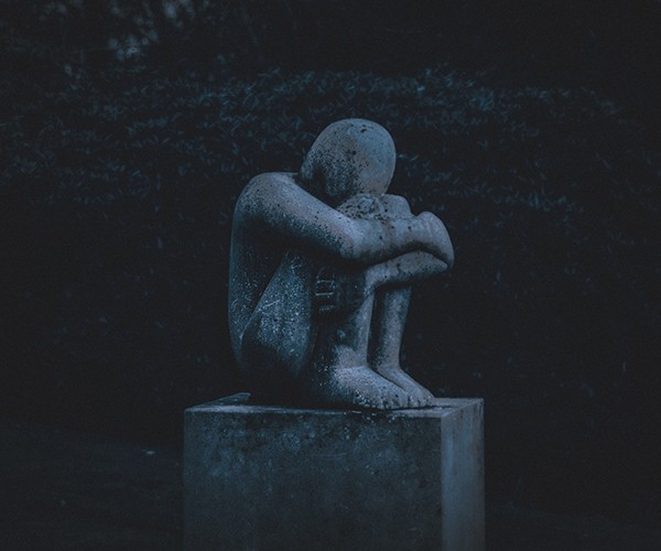 A stone statue of a faceless person with knees curled to chest, head down, arms wrapped around legs. Photo by K. Mitch Hodge on Unsplash