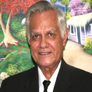 Dr. Alvelo wears a dark blazer white shirt and black tie while standing in front of a scenic painting of a cabin in a colorful woods setting.