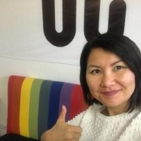 Guinara smiles giving a thumbs up sign wearning a beige colored sweater.  In the backdrop is a mini lounge covering of multi colored stripes in support of the pride community. 