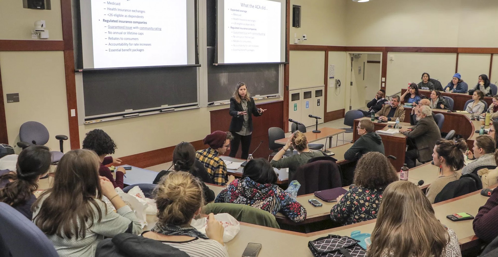 A graduate classroom full of students watch and listen to their female professor.