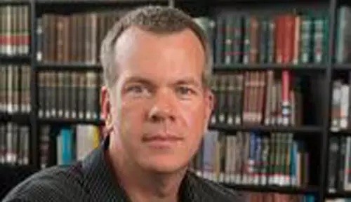 Dr. Epperson looks into camera he stands in front of a full bookshelf wearing a dark collared shirt. 