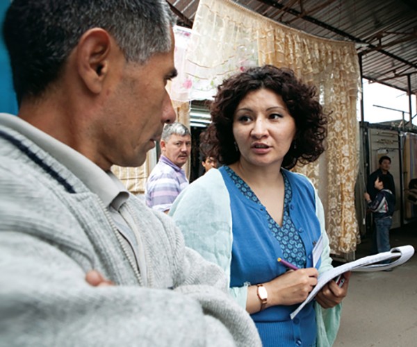 A woman writes on a notepad as she talks with a man in an outdoor marketplace.