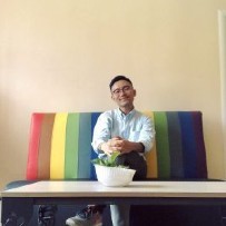 Yong Gun Lee sits crossed legged on a colorful multi striped lounge wearing a a light colored button down shirt and dark pants.  There is a coffee table in front of him with a plant inside of a white pot. 