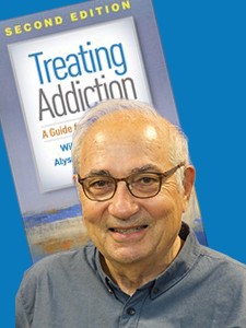 Allen wears dark rimmed eyewear and a grey shirt while his book cover is displayed in the backdrop