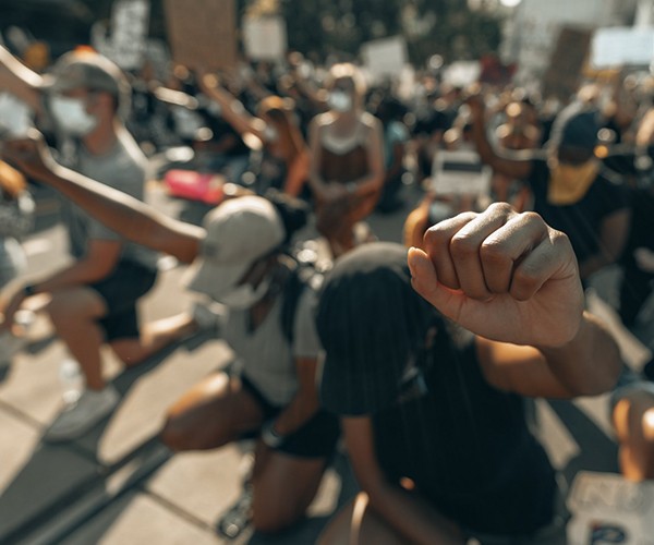 A group of people protesting for social justice kneel on the sidewalk with their fists raised.