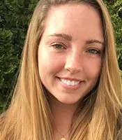 Molly Kowalski wears long flowing sandy blonde hair smiling into camera standing in front of green shrubbery.