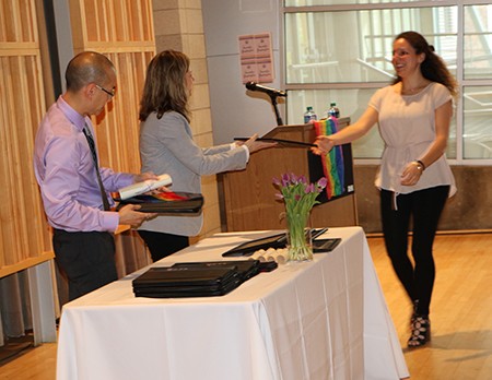 A woman walks up to recieve her diploma dressed in black pants and shoes with a tan top. She approaches the table where a man dressed in a lavendar button down shirt with dark pants. Another woman stands next to him wearing a grey jacket extending her hand towards the recipient.