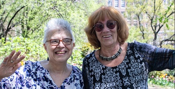 Professor Marion Riedel wears red rimmed eyeglasses and a blue and white multi colored blouse.  Professor Barbara Simon wears reddish brown hair dark sunglasses, a choker necklace and black an white multi pattern blouse 