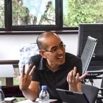 Dr. Wu wears a black t shirt while speaking with a smile and opened hands for expression.  His desk holds a bottled water and computer monitor.  His backdrop is a scenic dark paned window showing trees. 