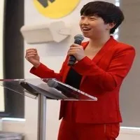 Ahsley wears a pixie haircut, a red blazer/pant suit witha black top underneath.  She holds a microphone at a plastic podium. 