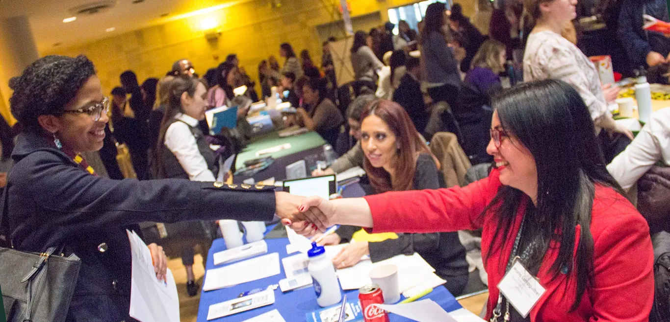 Two women shake hands over a table at a conference full of people. 