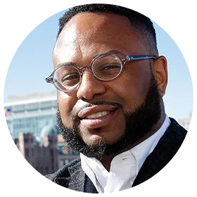Desmond smiles into camerw wearing dark rimmed eyelglasses, a white colored top and dark coat.