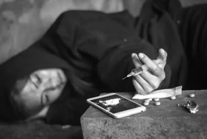 black and white photo of a woman lying on her side dressed in a dark hooded sweatshirt while holding a needle and drug paraphenilia strewn about. 
