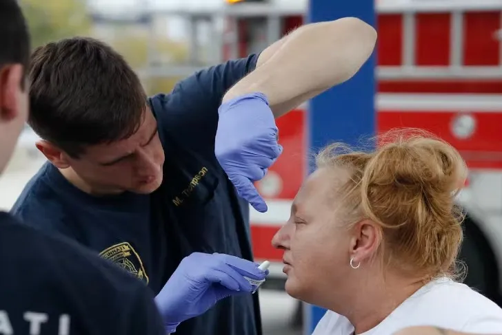 EMS team dressed in navy uniform and wearing purple gloves while tending to a woman dressed in a white top and gold earrings.  There are fire emergency vehicles parked in the background 