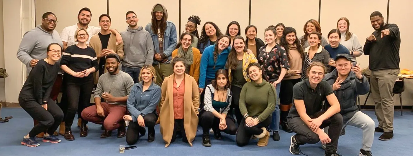 A group shot of various students pose in a blue carpeted room against beige walls.  