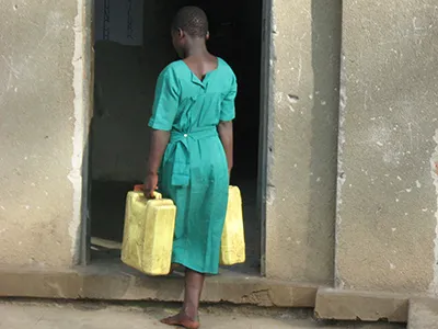 Ugandan youth wears a green cotton dress while carrying a yellow case in each hand.