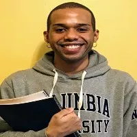 Westin Wallace stands in front of a yellow backdrop wearing a grey Columbia University hoodie sweatshirt.  He smiles and holds two books close to his chest. 