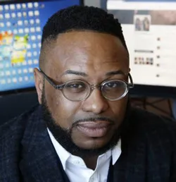 Up close image of Desmond Patton wearing a dark blazer, white collared shirt and dark wire framed eyeglass. He sits on front of two computer screens. 