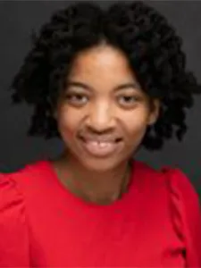Victoria Draper smiles at camera wearing a red blouse and dark natural twists in her hair. 
