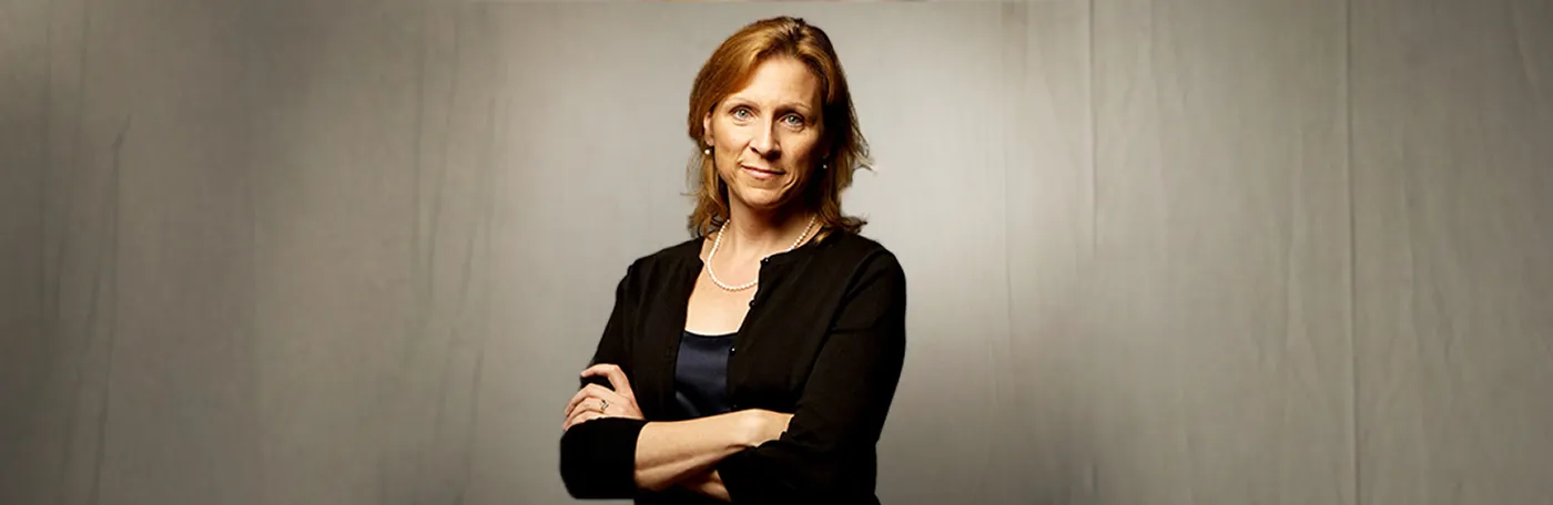 Dean Begg stands with arms folded dressed in a black top and sweater with a pearl necklace.