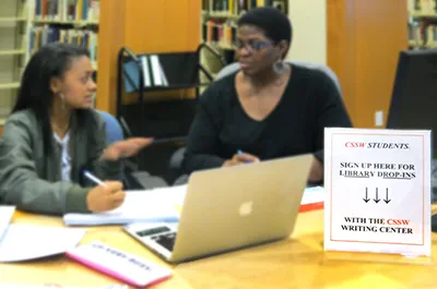 Two women seated at a table talk while using a laptop.