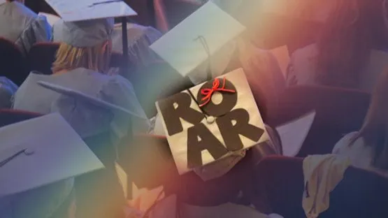 Overhead shot of multiple graduates wearing their graduate caps.  One particular cap adorns the letters ROAR which includes a red bow tie above the letter O.