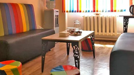 multi colored pride stripes adorn a mini dark leathered sofa. A mini water color is in the corner.  The window is covered in multi colored pride stripes above a radiator. There are multi colored pride striped mini ottomans positoned around a coffee table with cat iron decated legs. 