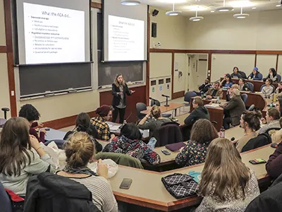 A graduate classroom full of students watch and listen to their female professor.