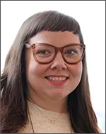 Rachel wears a smile and brown eyeglasses.  She has a beige sweater with a gold dainty chain on her neck and dark brown long hair. 